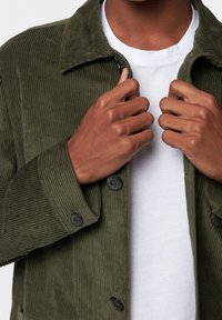 Oliwkowa sztruksowa kurtka z kołnierzykiem, z czarnymi guzikami, teksturowanymi prążkami i białą koszulką pod spodem.