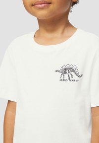 Witte katoenen T-shirt met een zwarte dino-skeletgrafiek en de tekst "#DINO TEAM UP" op de voorkant. Korte mouwen met een ronde hals.