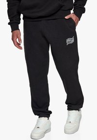 Sorte sweatpants med en blød tekstur, prydet med et hvidt grafisk logo på låret og elastiske manchetter ved anklerne. Sammen med hvide sneakers.