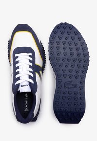 Lacoste tenisky v bílé syntetické svrchní části s námořnicky modrými semišovými detaily a žlutým pruhem. Gumová podrážka s trojúhelníkovým vzorem trakce.