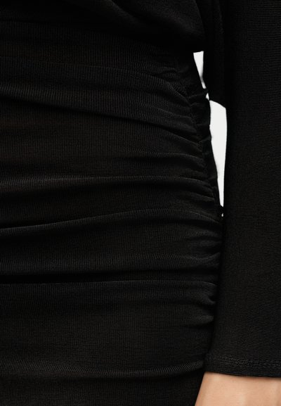 Μαύρο ριμπ ύφασμα με πλαϊνή φραδέλα, καλύπτοντας κορμό και χέρι, δείχνοντας την υφή και το συγκεντρωμένο ράψιμο σε κοντινό πλάνο.