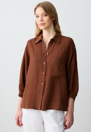 Camicia di lino marrone con maniche lunghe, chiusura a bottoni, taschino sul petto e vestibilità rilassata, abbinata a pantaloni bianchi. Tessuto morbido e finitura opaca.