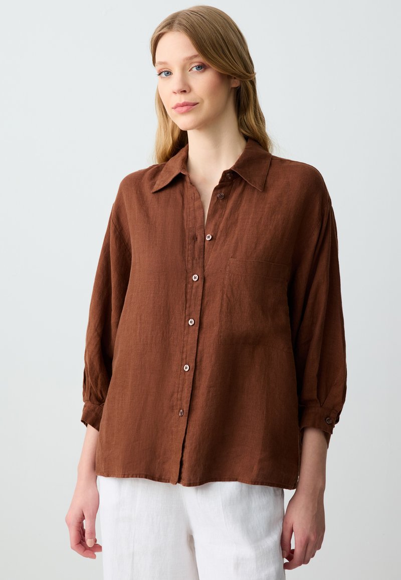 Camicia di lino marrone con maniche lunghe, chiusura a bottoni, taschino sul petto e vestibilità rilassata, abbinata a pantaloni bianchi. Tessuto morbido e finitura opaca.