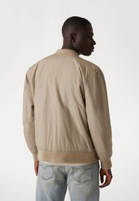 Veste bomber beige avec col et poignets côtelés, présentant une texture lisse et une taille élastique, portée sur un jean en denim clair.