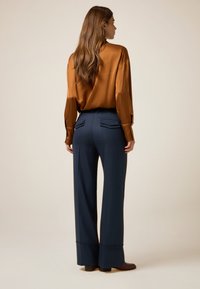 Blouse en satin marron à manches longues et poignets boutonnés, associée à un pantalon large bleu marine avec poches arrières et un discret détail d'ourlet.