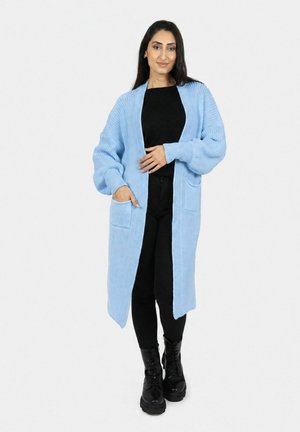 Vrouw draagt een lichtblauwe lange gebreide cardigan over een zwart topje, zwarte broek en zwarte veterlaarzen, staand tegen een effen achtergrond.