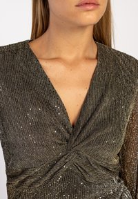 Robe métallique verte avec un décolleté en V profond, tissu texturé avec des rayures verticales et de subtils sequins, mettant en valeur le design noué à l'avant.