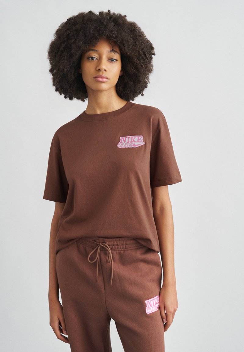 Καφέ βαμβακερό T-shirt με ροζ κεντητό λογότυπο "NIKE CLUB" στο αριστερό στήθος, συνδυασμένο με καφέ παντελόνι φόρμας με κορδόνι σετ.