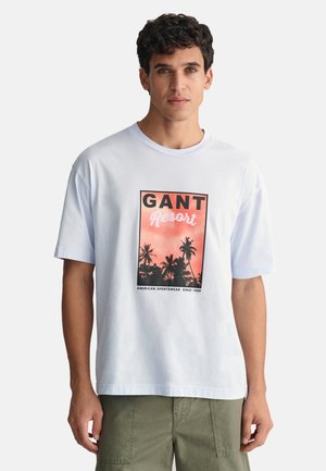 Camiseta de algodón blanca con un gráfico frontal que presenta el texto "GANT Resort" sobre un diseño de palmera en rosa y negro. Manga corta y cuello redondo.
