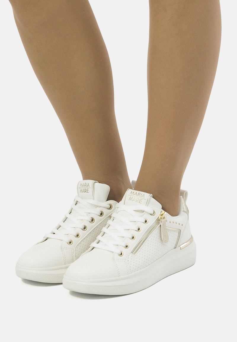 Baskets blanches avec des côtés perforés, accents en matériel doré et design à lacets. Elles possèdent une fermeture éclair sur le côté et une semelle rembourrée pour plus de confort.