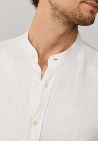 Hombre que lleva una camisa de lino blanca con cuello mandarín, abotonada cerca del cuello, mostrando parte de la mandíbula y el cuello.