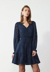 Robe bleu marine avec de longues manches transparentes, décolleté en V et taille smockée. Dispose d'une jupe à plusieurs couches et d'une texture florale pour plus de détails.