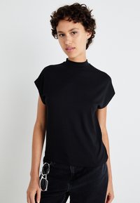 Svart t-shirt med kort ärm och hög krage, tillverkad i mjukt material. Har en avslappnad passform, kombineras med mörka denimbyxor och solglasögon.