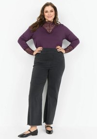 Kobieta stoi z rękami na biodrach, ubrana w fioletową bluzkę z koronkowym wykończeniem, czarne dżinsy z wysokim stanem i szeroką nogawką oraz czarne buty Mary Jane.