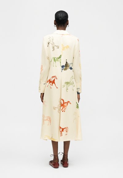 Γυναίκα με σκουρόχρωμο δέρμα και μαλλιά μαζεμένα σε κότσο, φοράει μακρύ κρεμ παλτό με πολύχρωμες εκτυπώσεις αλόγων και καφέ δερμάτινα παπούτσια με κορδόνια, στέκεται μπροστά σε απλό φόντο.