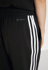 Sorte træningsbukser med elastik i taljen, der har tre hvide lodrette striber og et lille "AEROREADY"-logo på siden.