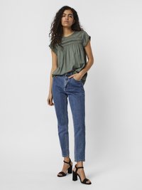 Blouse olive verte à manches courtes avec des détails de smocks, associée à un jean en denim bleu taille haute et des sandales à talons carrés noires.