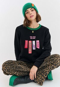 Černá mikina s barevnými textovými nášivkami ("LÁSKA," "ROVNOVÁHA," "RADOST," "RIZIKO") na přední straně, spojená s kalhotami s leopardím potiskem a zelenými platformovými botami.