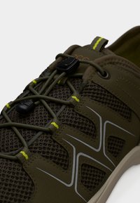 Κοντινό πλάνο αθλητικού παπουτσιού σε ελιά πράσινο χρώμα με υφαντό ύφασμα, ελαστικά κορδόνια τύπου bungee και ανακλαστικές λωρίδες στο πλάι.