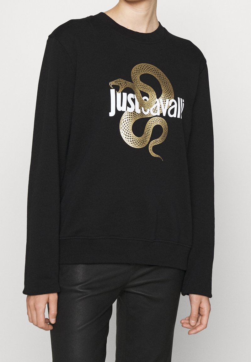 černá mikina s zlatým grafikou hada ovíjejícího se kolem bílé textu "Just Cavalli", nošená s černými kalhotami.