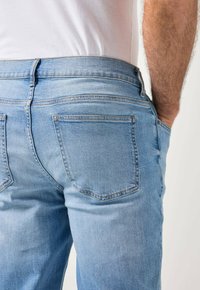 Mies, jolla on vaaleansiniset farkut ja valkoinen paita, seisoo toisella kädellä farkkujen takataskussa.