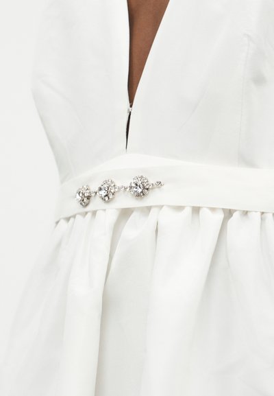 Λευκό φόρεμα με βαθύ ντεκολτέ σε σχήμα V και εφαρμοστό ζωνάρι στη μέση, διακοσμημένο με τρία στρογγυλά κρυσταλλικά στολίδια πάνω από την πτυχωτή φούστα.