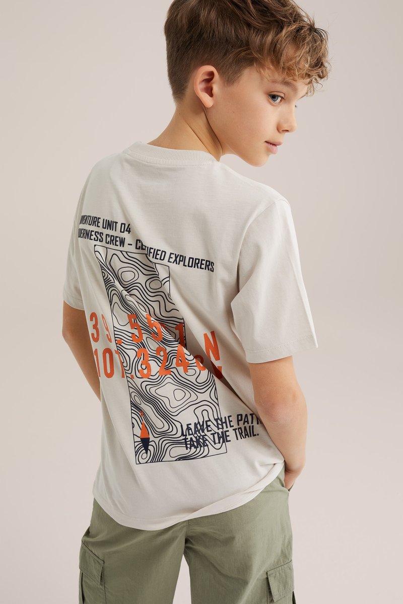 Pois lokkis juustega, kes kannab valget T-särki musta ja oranži topograafilise kaardi kujundusega ning oliivrohelisi lahtisi lühikesi pükse, pööratud küljele.