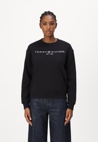 Sudadera negra con cuello redondo, que presenta el logotipo en blanco "TOMMY HILFIGER EST. 1985". Tejido suave y ajuste relajado, combinada con jeans anchos azules.