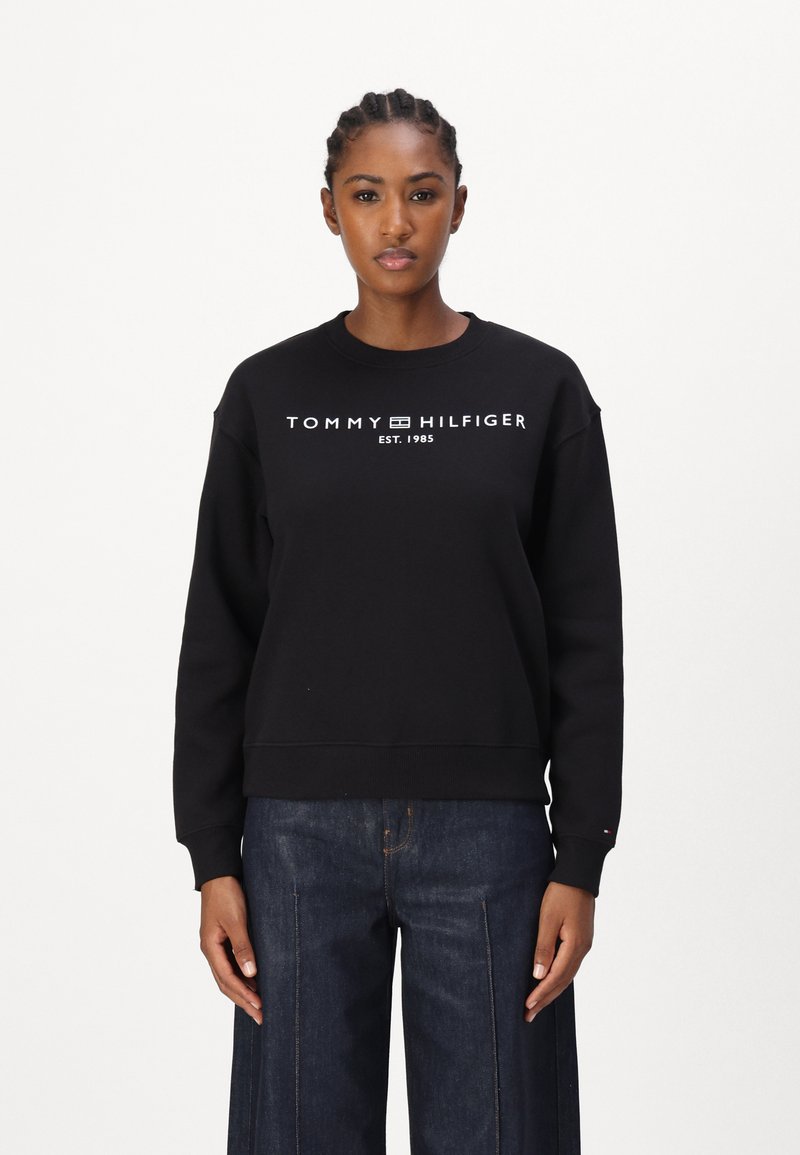 Sudadera negra con cuello redondo, que presenta el logotipo en blanco "TOMMY HILFIGER EST. 1985". Tejido suave y ajuste relajado, combinada con jeans anchos azules.