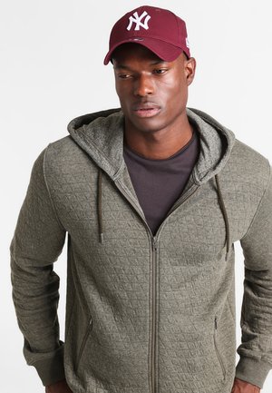 Olivgrüner, gesteppten Reißverschluss-Hoodie mit einer grau gefütterten Kapuze und verstellbaren Kordeln. Getragen über einem dunklen T-Shirt, kombiniert mit einer kastanienbraunen Mütze.