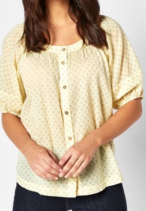 Femme portant un chemisier jaune clair à manches bouffantes avec de petits motifs circulaires, boutonné sur le devant, associé à un jean foncé.