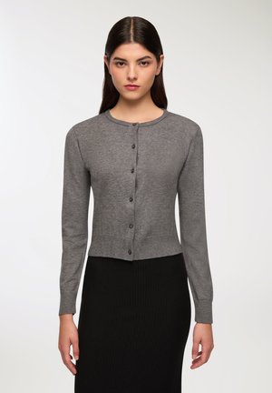 Cardigan grigio con collo rotondo, maniche lunghe e chiusura con bottoni. Vita a coste. Texture morbida. Abbinato a una gonna nera a coste.