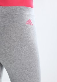 Detailní záběr na šedé sportovní legíny s růžovým logem Adidas na horním stehně, v kombinaci s jasně růžovým topem.