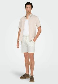 Camisa corta beige claro con botones, camiseta sin mangas blanca, pantalones cortos blancos texturizados y zapatos de gamuza marrón con cordones, todo sobre un fondo neutro.