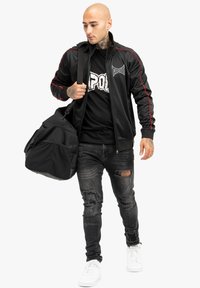 Chaqueta de chándal negra con acentos rojos, camiseta negra de TAPOUT, jeans negros desgastados, zapatillas blancas, llevando una gran bolsa de deporte negra.