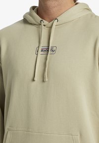 Sudadera beige con cordones, que cuenta con un bolsillo delantero y un parche de logo azul. Fabricada con un tejido suave y texturizado, adecuada para el uso casual.