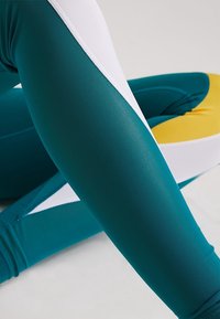 Legginsy w kolorze turkusowym i białym wykonane z elastycznej tkaniny; cechują się gładką teksturą oraz wzorem w blokach kolorystycznych z kontrastowym żółtym akcentem.