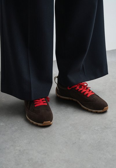 Ténis de camurça castanho com atacadores vermelhos e solas de borracha castanha, combinados com calças escuras de riscas. Os ténis têm um perfil baixo e detalhes texturizados.