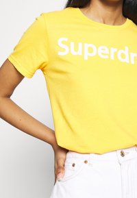 Camiseta de algodón amarilla con mangas cortas que presenta el logo "Superdry" en texto blanco en negrita. Tejido ligero, cuello redondo clásico.