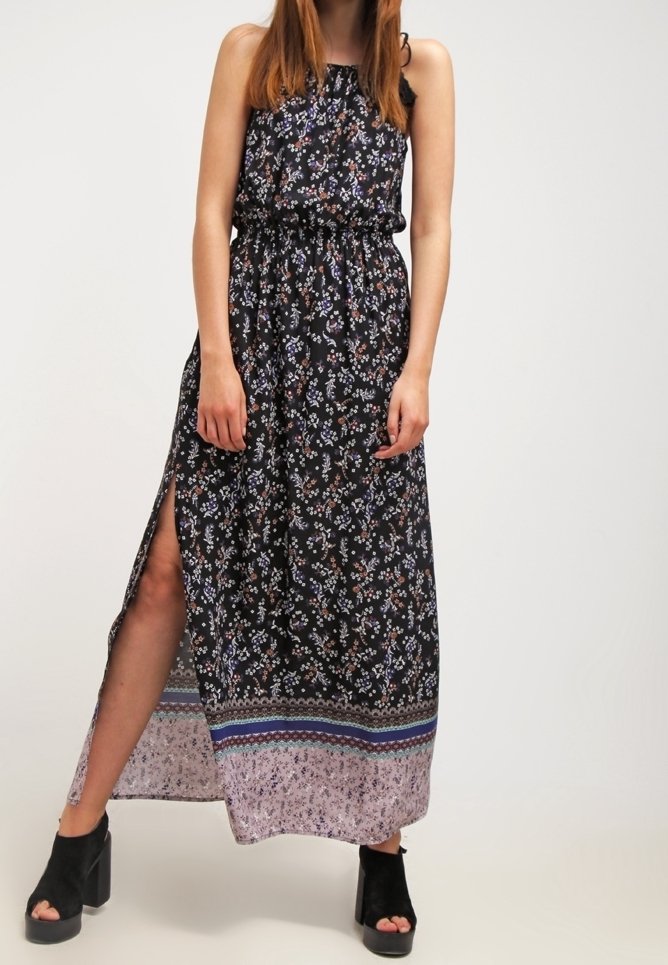 Femme portant une robe longue noire à fleurs sans manches avec une fente latérale et des talons noirs ouverts à plateforme, debout devant un fond uni.