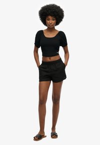 Zwarte cropped top met korte pofmouwen, van textuurstof; gecombineerd met zwarte shorts met elastische tailleband. Model draagt zwarte slide sandalen.