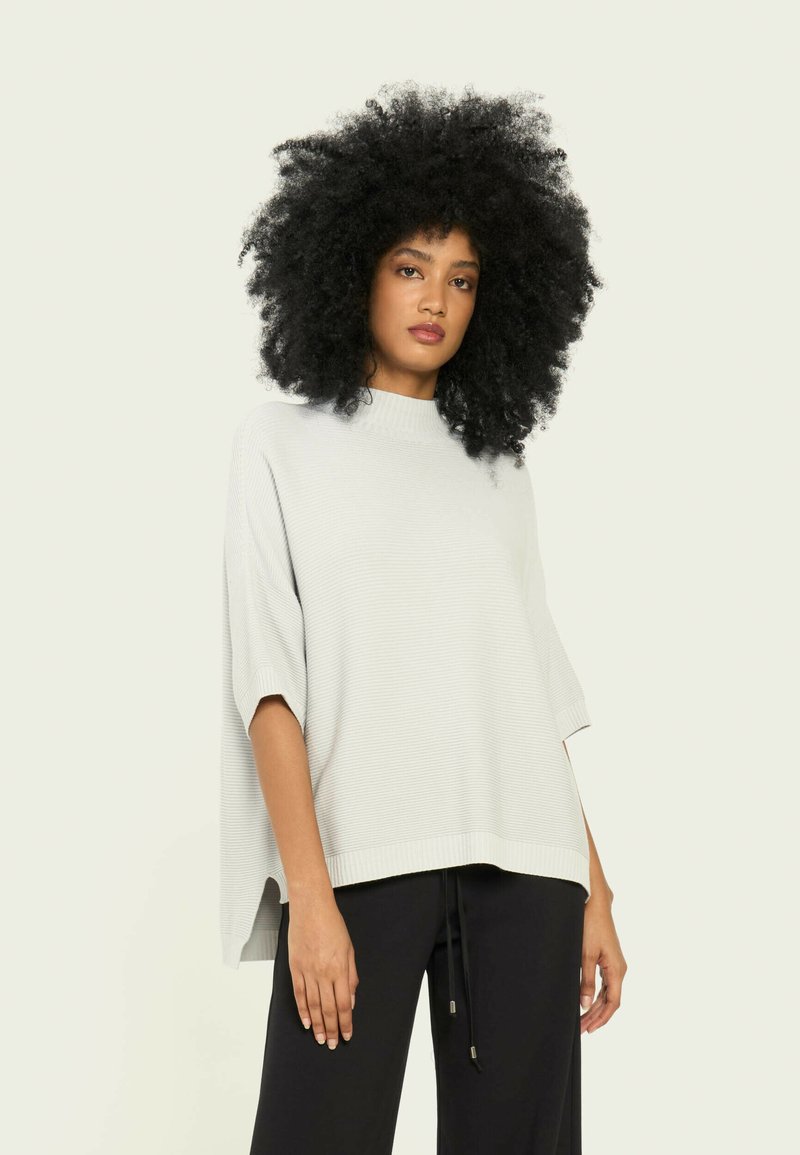 Pulover oversized de culoare gri deschis, cu guler înalt, textură ribbed și mâneci scurte, asortat cu pantaloni închiși la culoare.