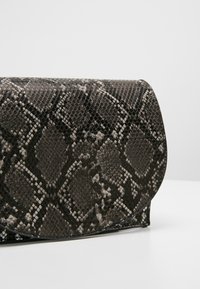 Bolso de mano con un patrón de piel de serpiente negro y gris y borde superior curvado, apoyado sobre una superficie blanca.