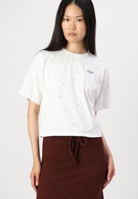 Hvit kortermet t-skjorte med små steiner, prydet med "Pepe Jeans London"-logoen trykt på. Kombinert med et brunt skjørt med snøring.
