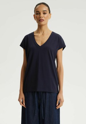 Kobieta ubrana w granatowy top z krótkim rękawem i dekoltem w serek oraz luźne, ciemnoniebieskie dżinsowe spodnie, stojąca na prostym jasnym tle.