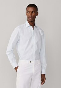 Homme portant une chemise blanche à manches longues à boutons avec un motif à carreaux bleu clair et un pantalon blanc, debout avec une main dans la poche.
