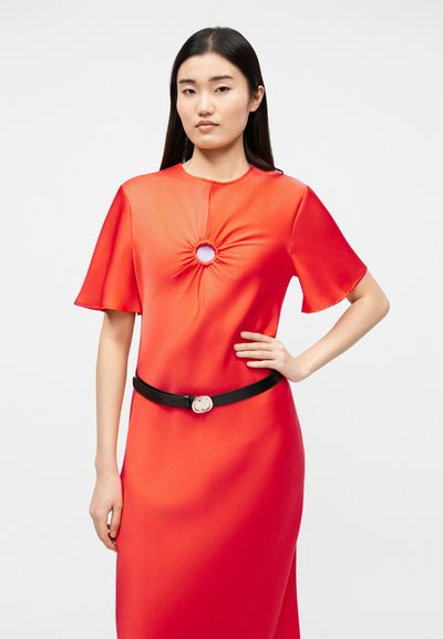 Γυναίκα με μακριά μαύρα μαλλιά που φοράει φωτεινό κόκκινο φόρεμα με μανίκια που πετούν και ραμμένη λεπτομέρεια με άνοιγμα στο στήθος, στερεωμένο με μαύρη ζώνη.