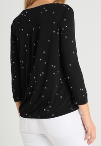 Zwarte blouse met lange mouwen en metallic sterrenpatronen, met een ronde halslijn en een geplooid detail aan de achterkant, gedragen met witte broek.