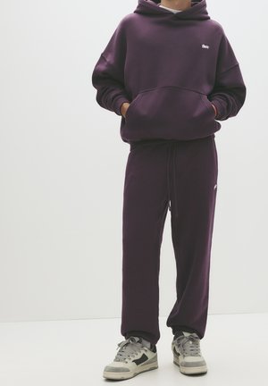 Lilla hættetrøje med en frontlomme og broderet logo, parret med matchende lilla joggers. Båret med grå sneakers. Glat tekstur.