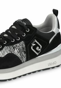 Des baskets noires en daim avec un panneau latéral en relief argenté et un accent argenté brillant. Elles sont équipées de lacets noirs et d'une semelle épaisse blanche.
